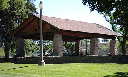 lawn area foreground with rock seat wall, lamp post and large picnic shelter with timber beams and rock columns and trees on sides and in background
