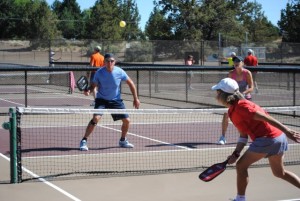 Pickleball players at Pine Nursery Park courts. Foreground includes 3 pickleball players actively playing the game. The background includes 5 others playing and talking at the Pine Nursery pickleball courts.