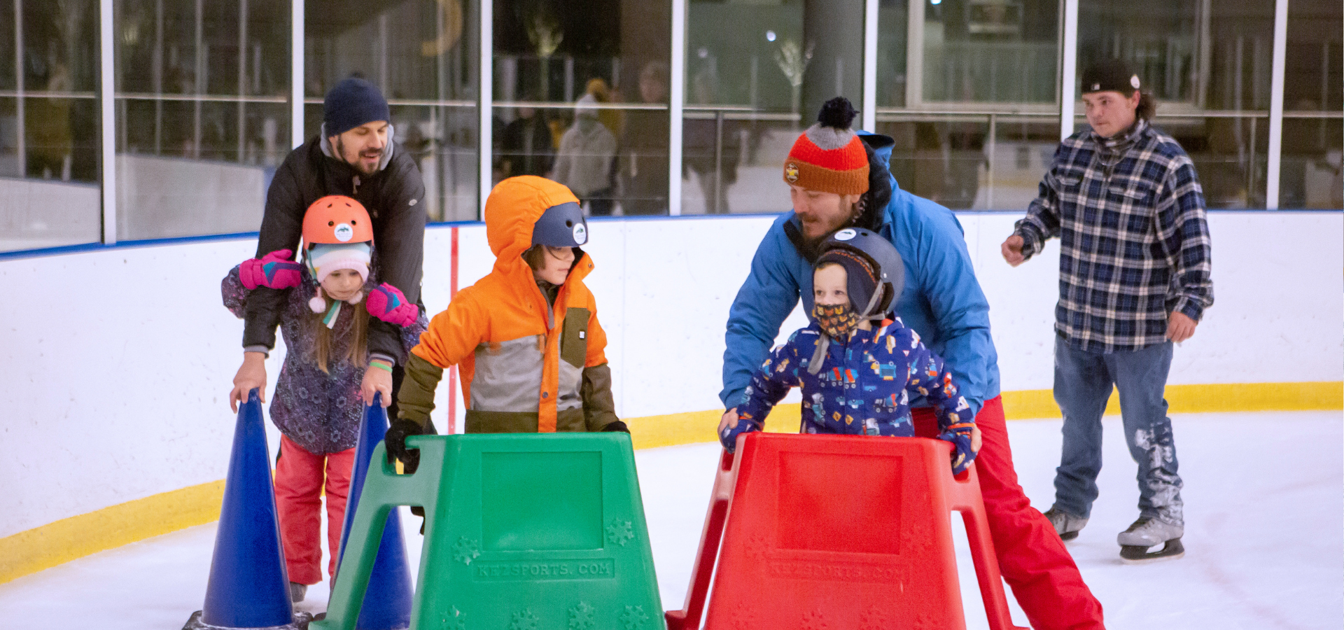 At the Pavilion, a group of children and adults use skate assisting items to make their way on the ice.