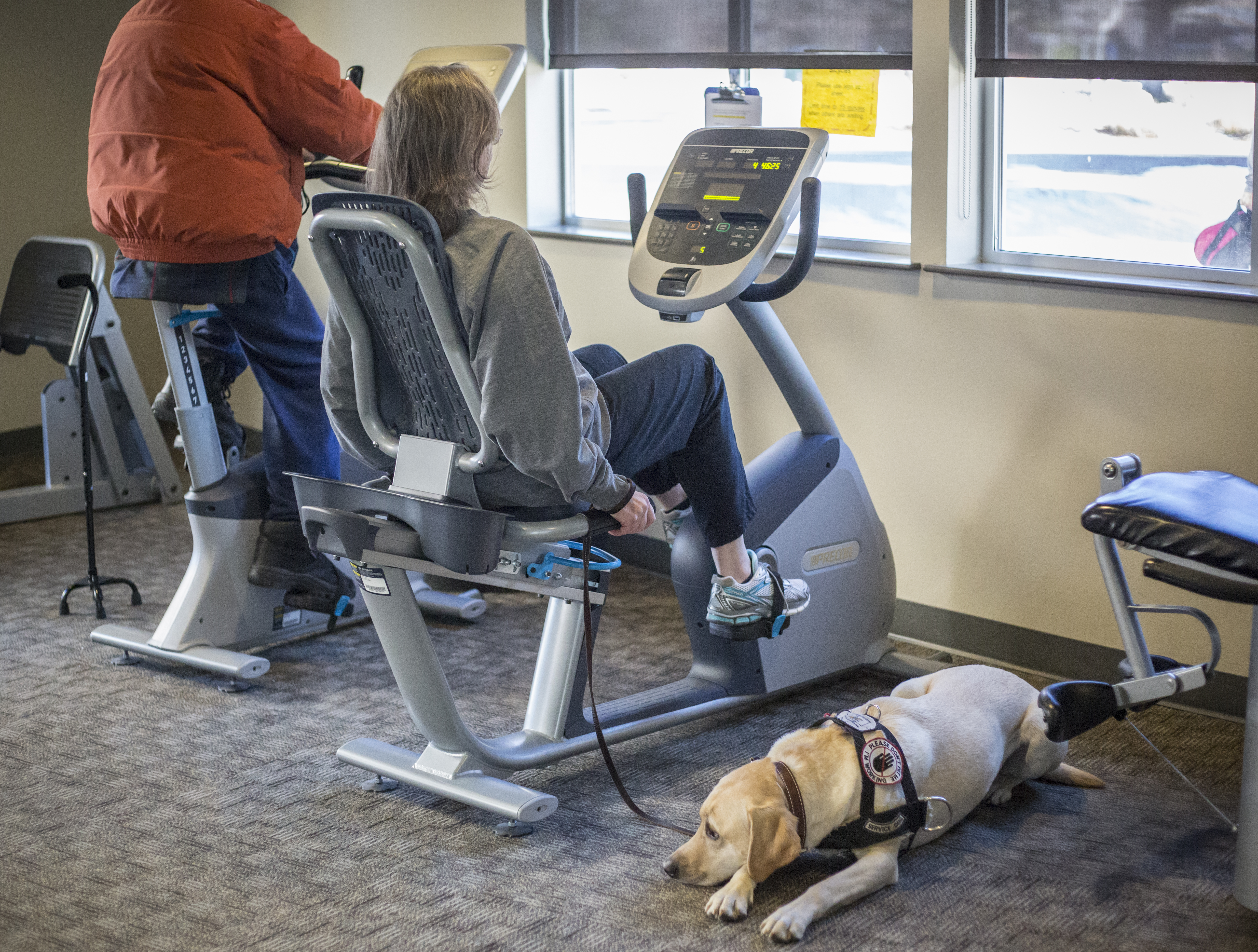 A dog rests while a woman operates a cycling machine at the Social activity at Bend Senior Center.