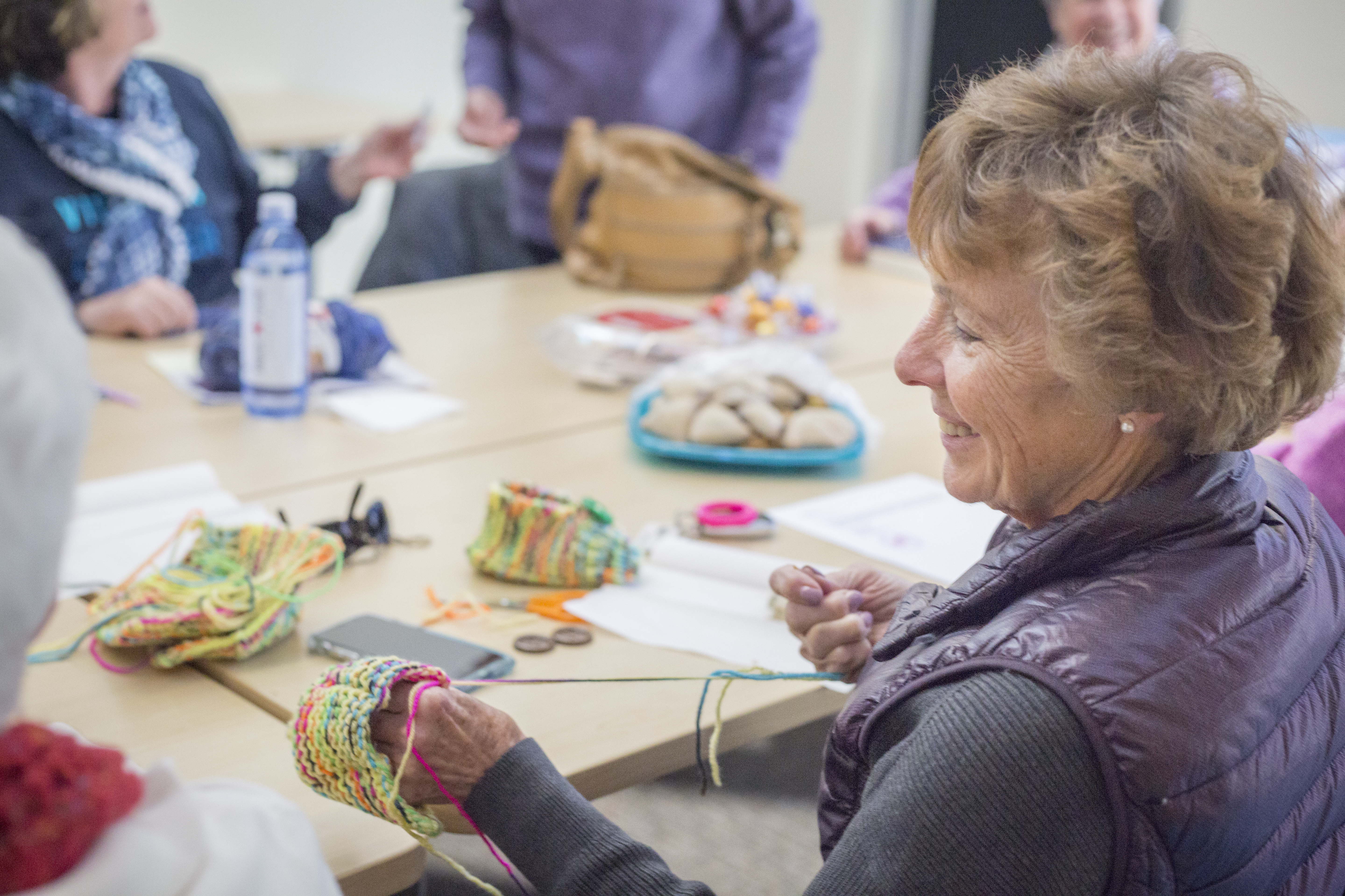 A woman smiles amongst a group of adults engaging in a social activity involving yarn.