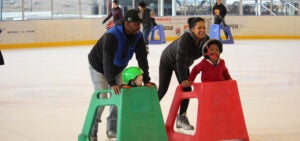 A mother and father skating with their two children, both using skate assist devices at the Pavilion
