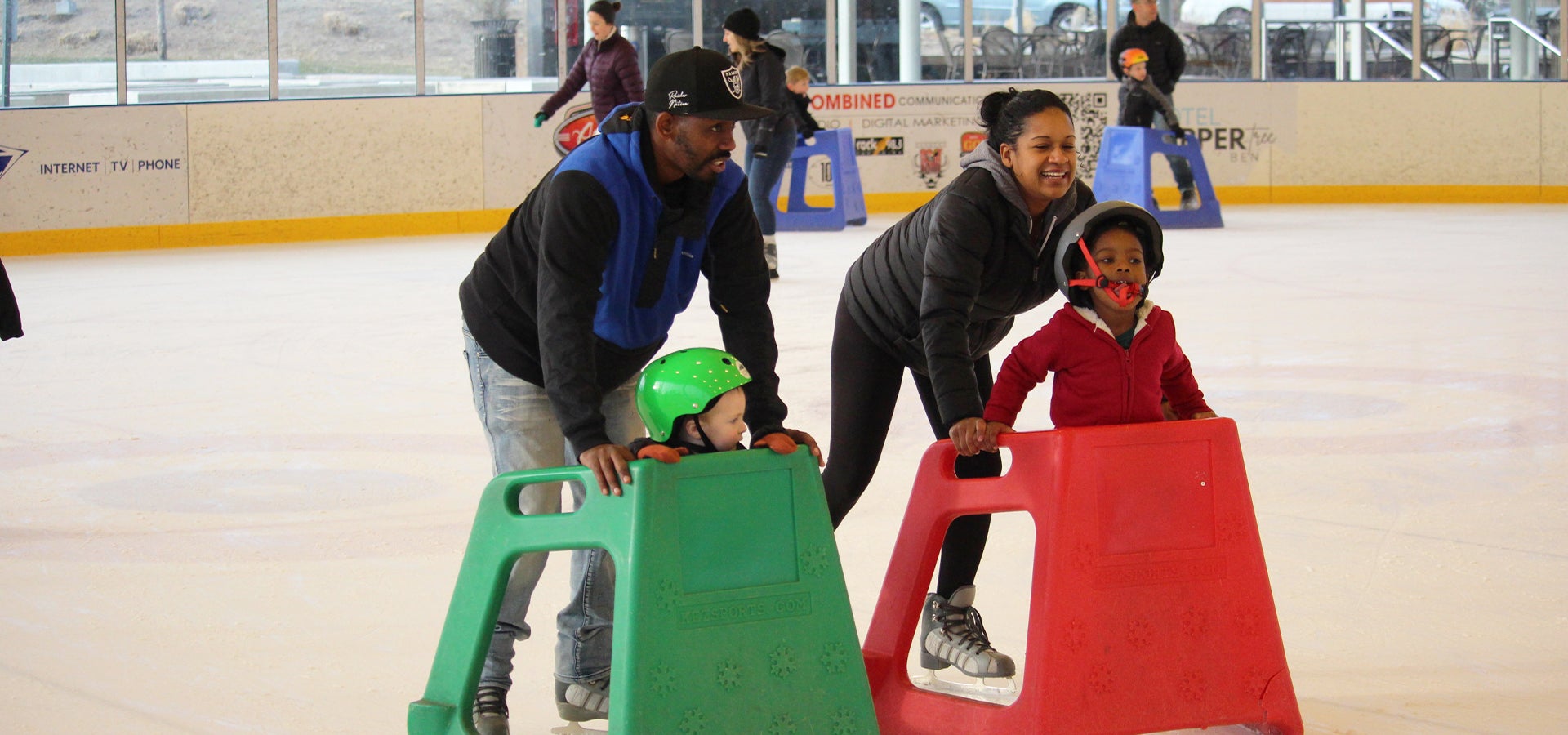 A mother and father skating with their two children, both using skate assist devices at the Pavilion