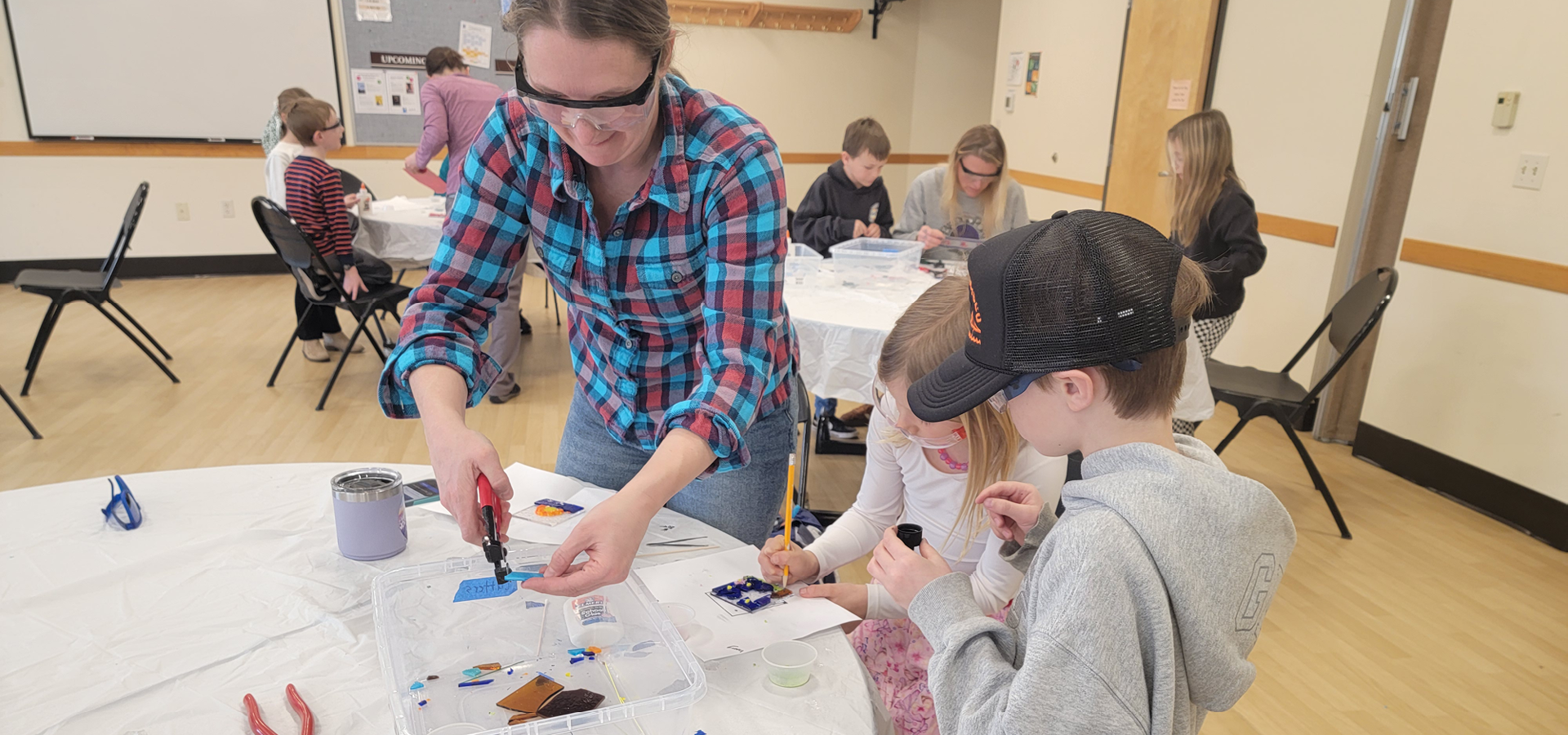 Group of adults and children work on glass arts projects on tables throughout a room