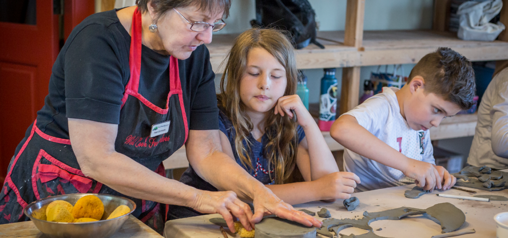 An instructor guides a child in a pottery class while another child works on a piece.