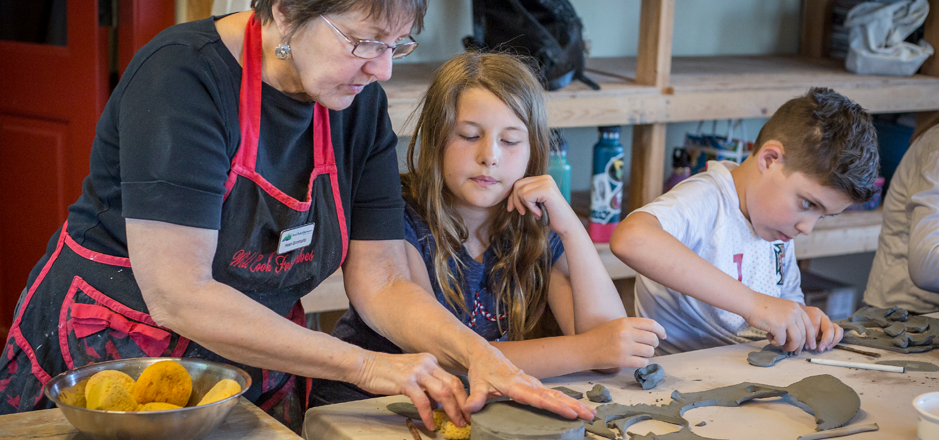 An instructor guides a child in a pottery class while another child works on a piece.