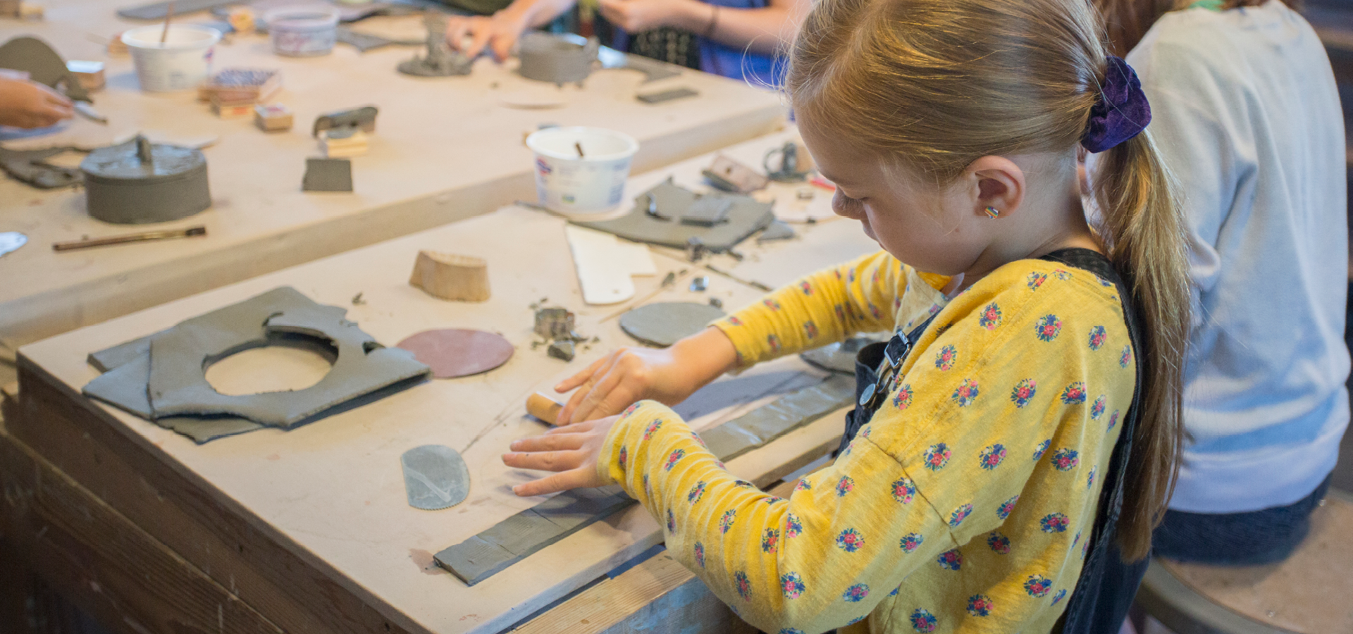 A small child works on a handbuilt pottery piece on a table scattered with pieces of clay
