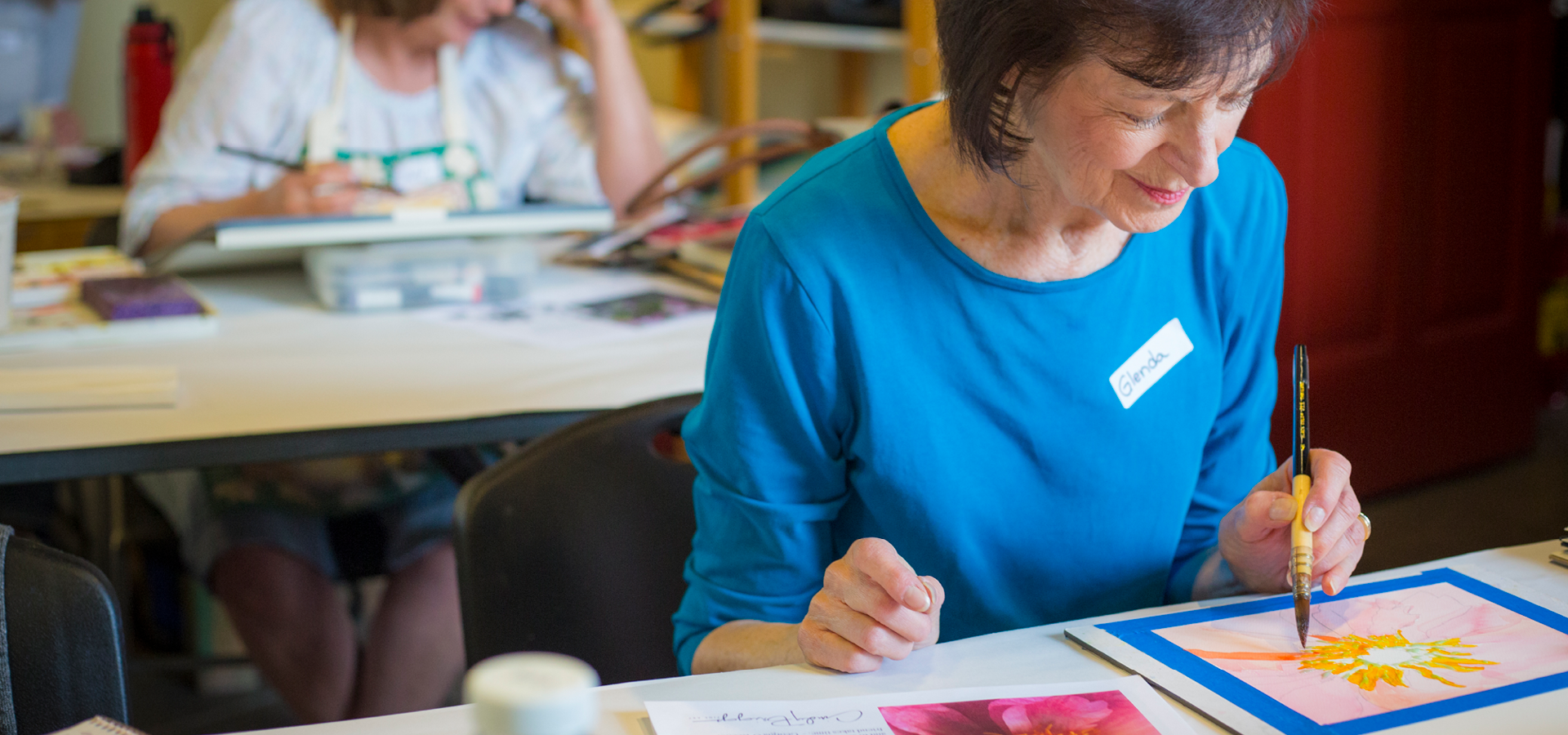 A woman smiles and paints a small floral painting in watercolor class