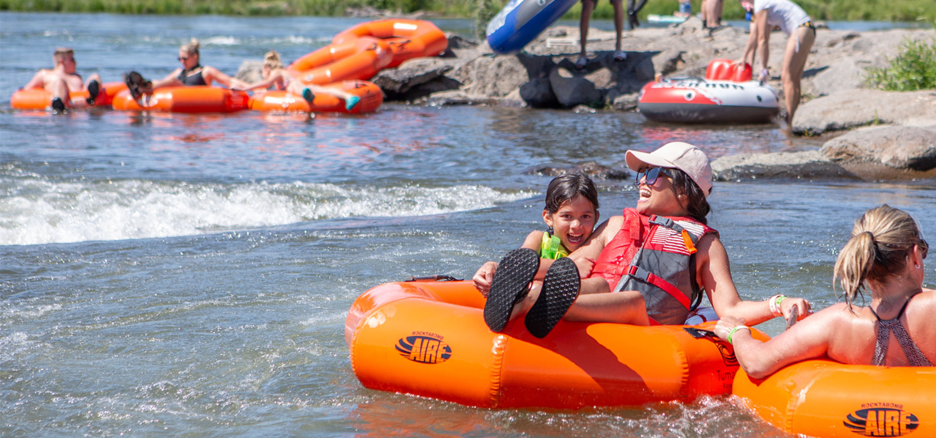 A child and gaurdian wearing lifevests and water shoes smile as they float down the river on a river tube.
