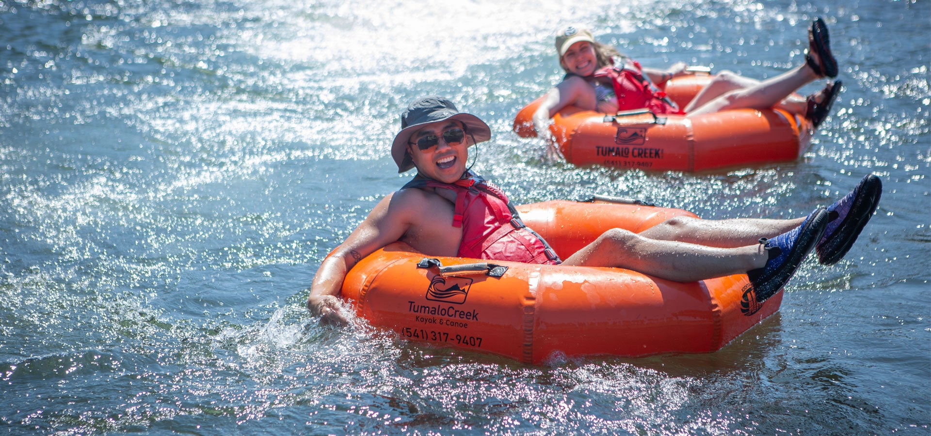 Two adults smile at the camera while enjoying floating the river in tubes with life jakcets and water shoes.