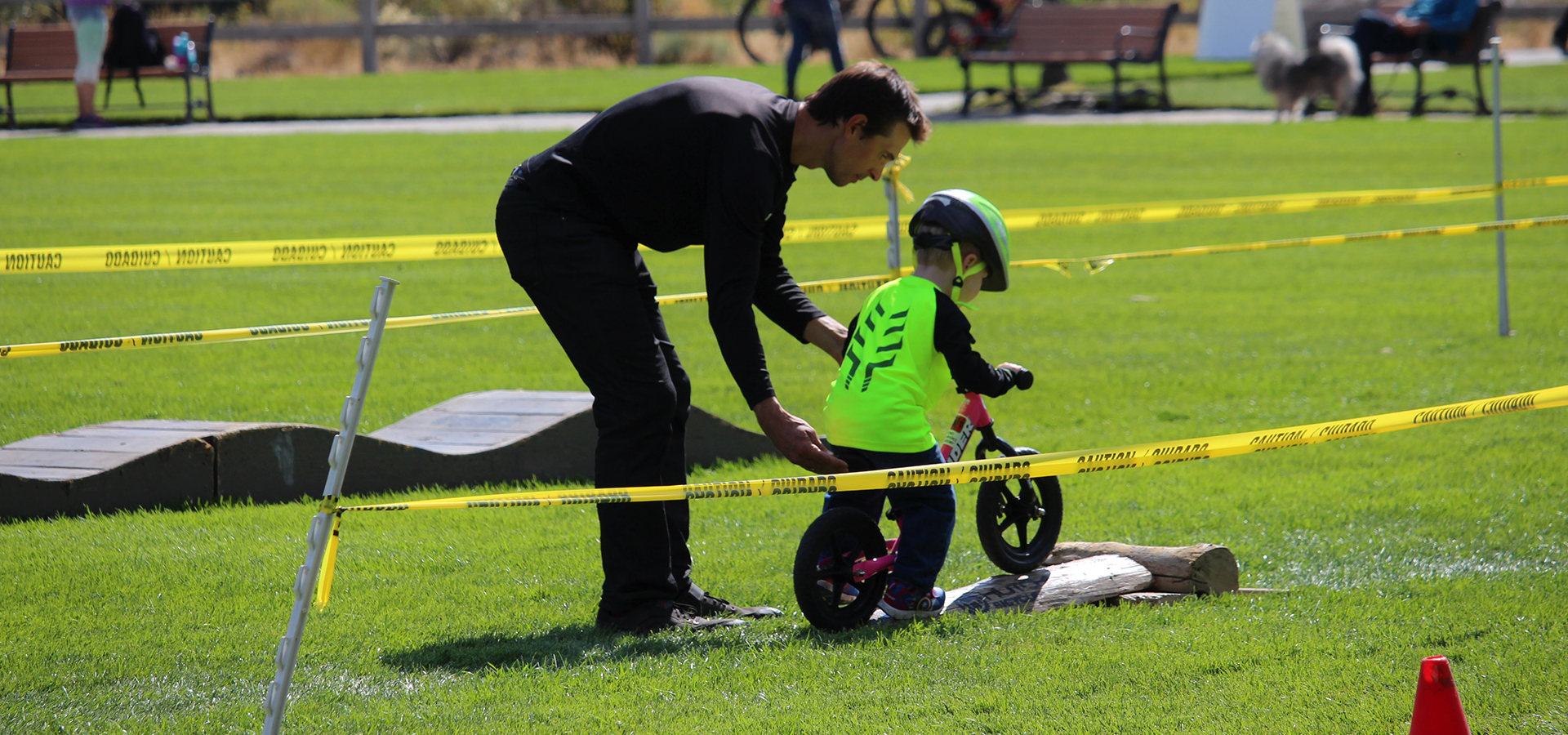 A little boy riding a bike over an obstacle with an adult helping.
