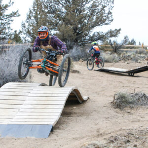 a cyclist on an adaptive bike gets some air as he rides across a wood ramp obstacle and another cyclists rides in the background