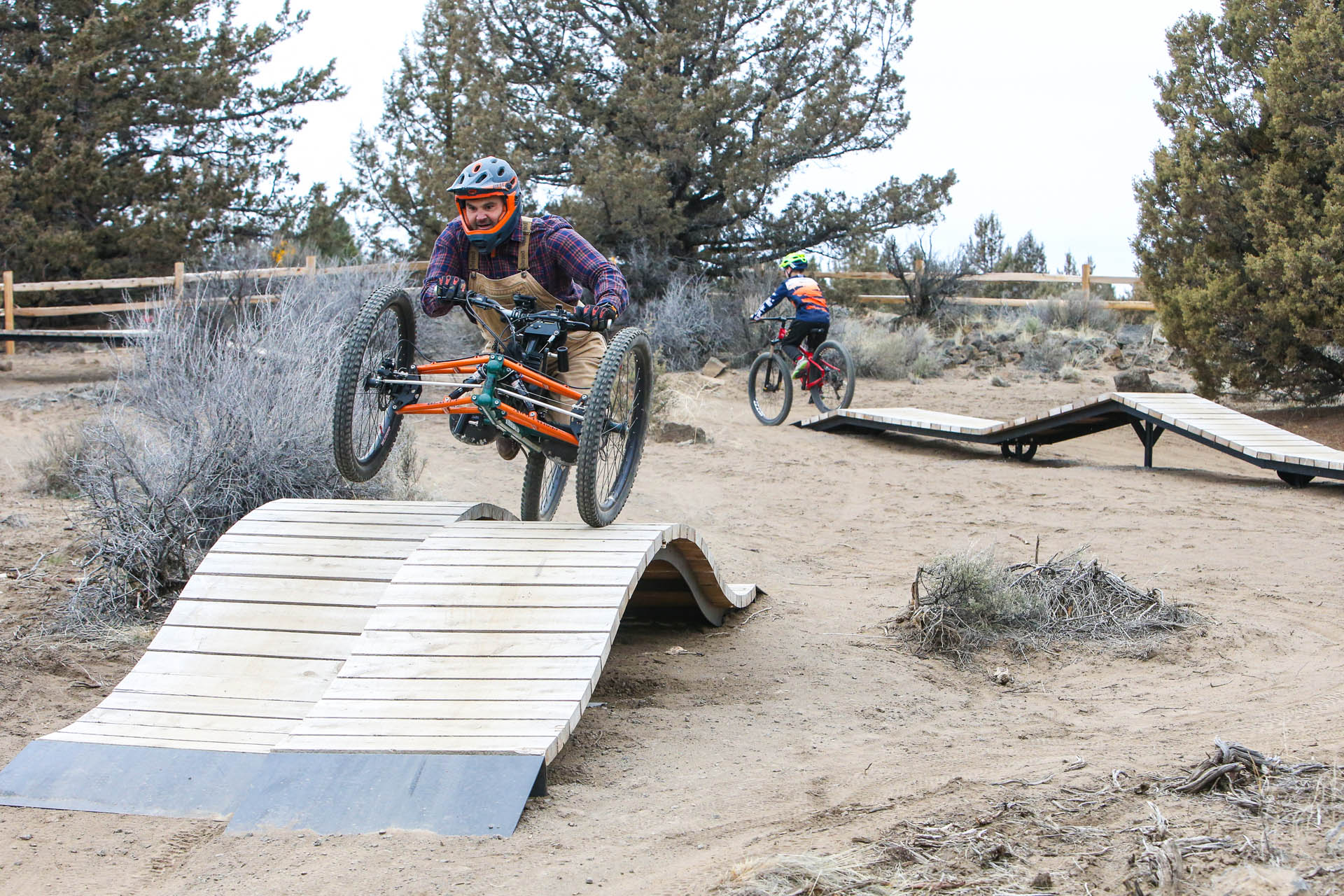 a cyclist on an adaptive bike gets some air as he rides across a wood ramp obstacle and another cyclists rides in the background