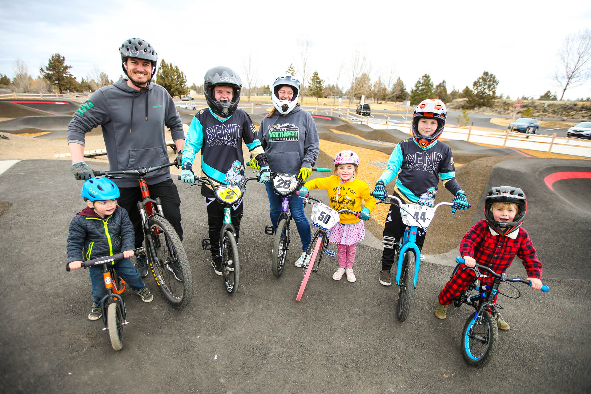 a group of bike riders of variety of ages and sizes pose in front of the pump track with their bikes