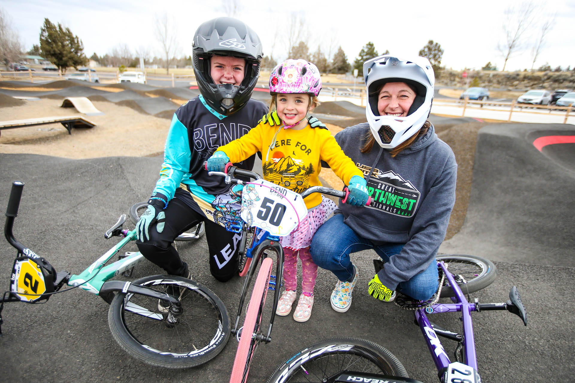 two children and one adult smile in front of the pump track with their bikes