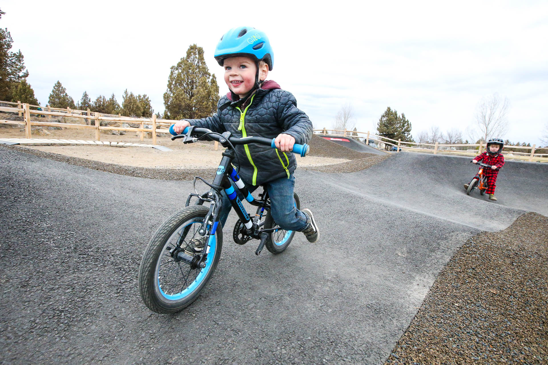 a child on a bike rides on the pump track with another child in the background