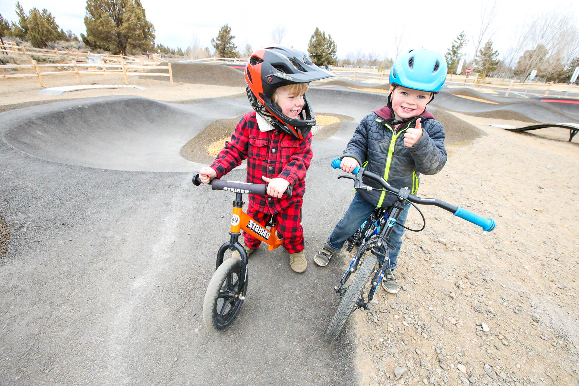 two young children on bikes pose in front of pump track, one is giving a thumbs up