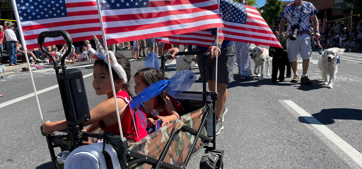 two children ride in a American flag decorated wagon with parade patrons in background