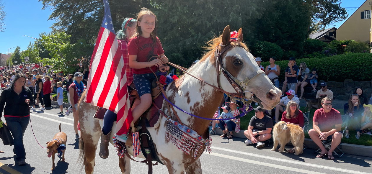 two children ride an American flag decorated horse through crowded parade route