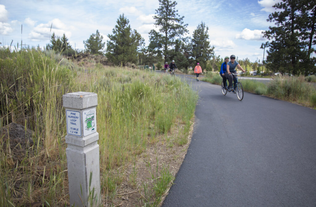 Paved path with trail sign in the grass at Pine Nursery park with bikers in the background