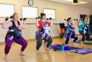 Three patrons workout lift weights and squat while wearing babies in harnesses.