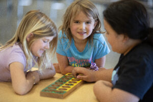 Three children play a tabletop game together while one smiles at the camera.